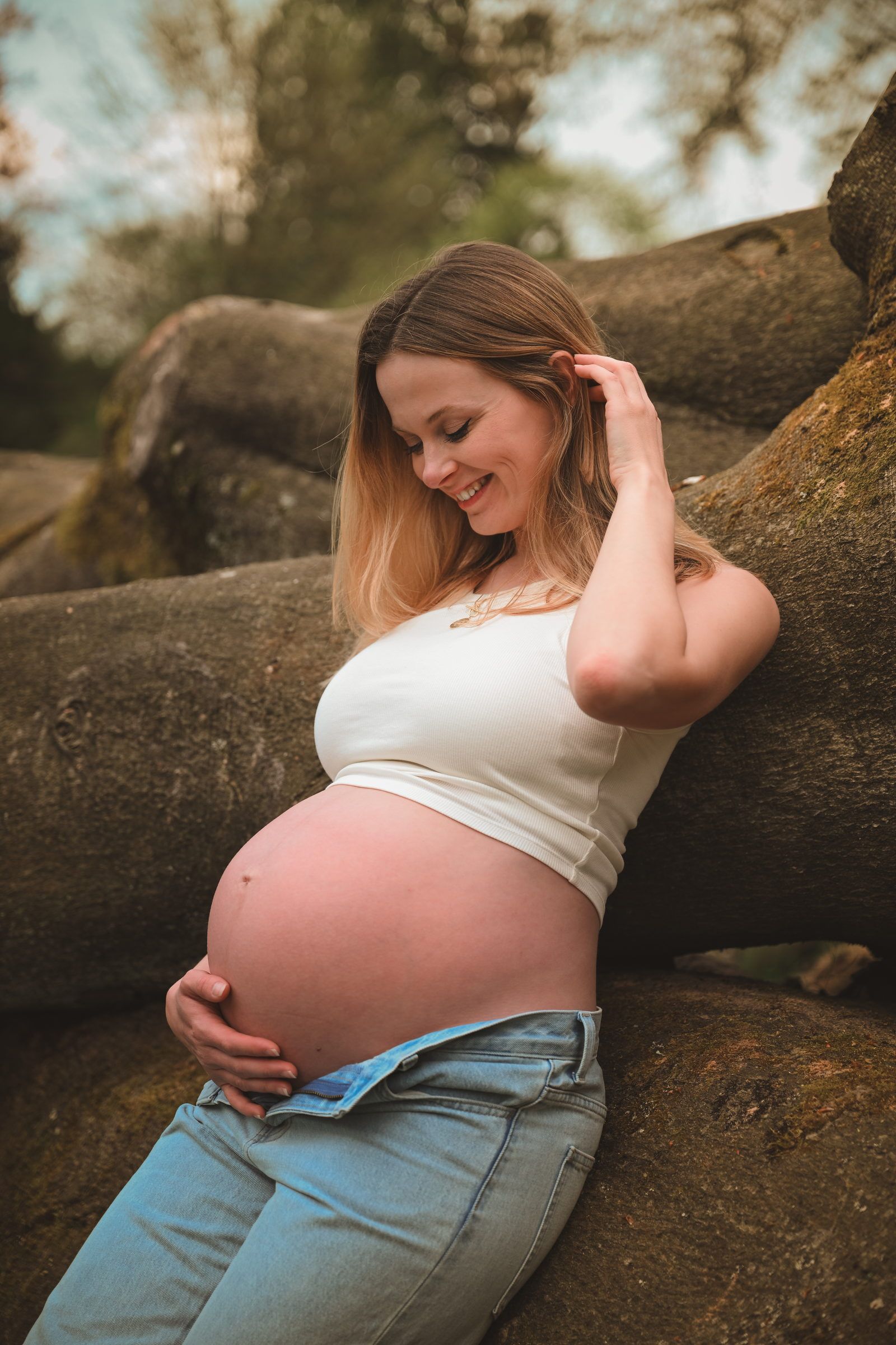 Femme enceinte adossée à des racines, qui sourit en regardant son ventre