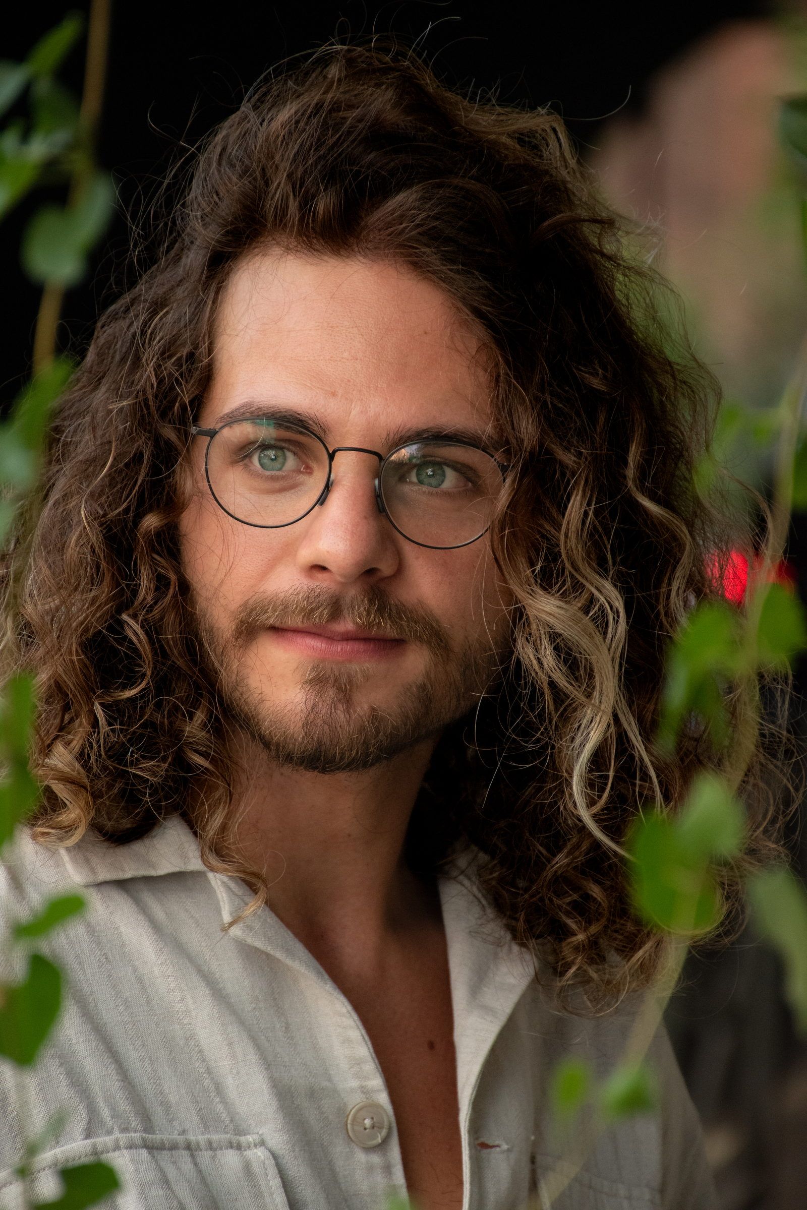 Homme aux longs cheveux bouclés et aux lunettes rondes, qui regarde l’appareil à travers un feuillage
